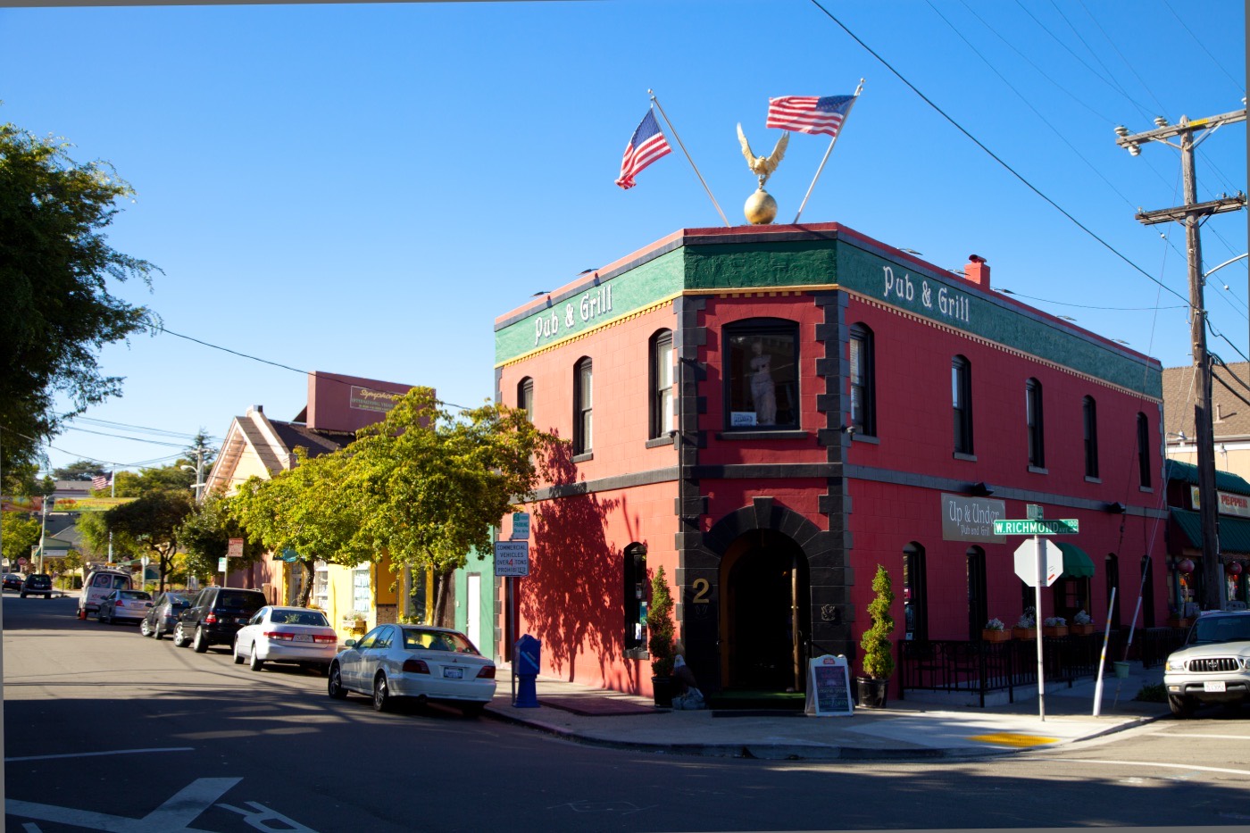 A red corner building housing the Up & Under Pub & Grill in Point Richmond's historic district, with American flags on the roofline and parked cars along the street.